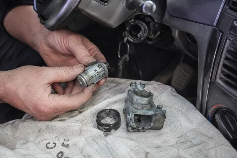 Hands holding a removed ignition cylinder beside a car steering column, with the housing and small parts on a cloth. — Ignition Lock Repair, Welland
